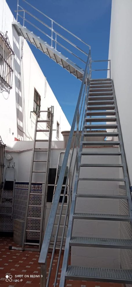 Escalera metálica con barandillas, subiendo hacia una plataforma sobre un fondo azul.