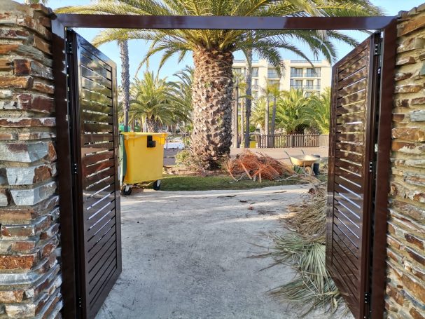 Puertas abiertas hacia un jardín con palmera y un contenedor amarillo al fondo.