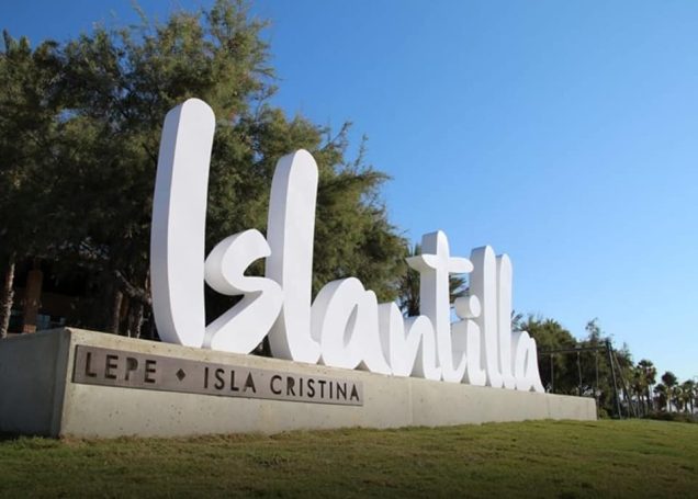 Letrero grande y blanco que dice "Islantilla" con un fondo de cielo azul y árboles.